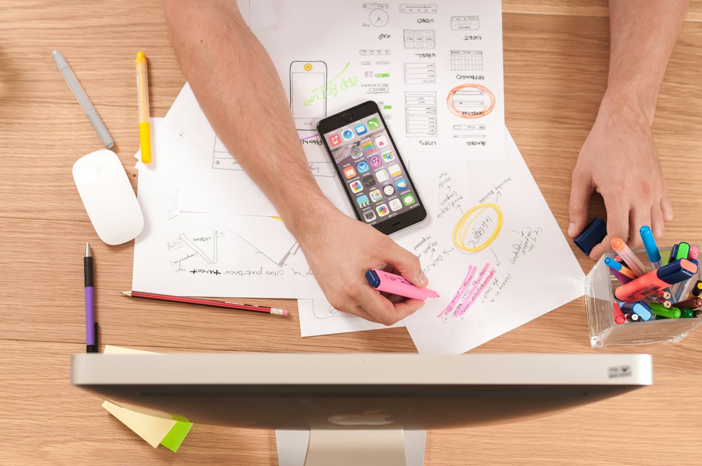 Hands highlighting notes on a wooden desk with design sketches and a smartphone.